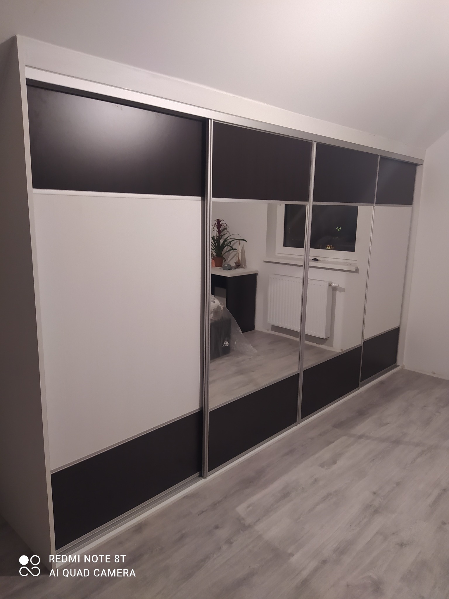 Duża szafa przesuwna z lustrzanymi drzwiami, dzielona na trzy poziome pasy: czarny, biały i czarny. W lustrach odbija się pokój z grzejnikiem i biurkiem.