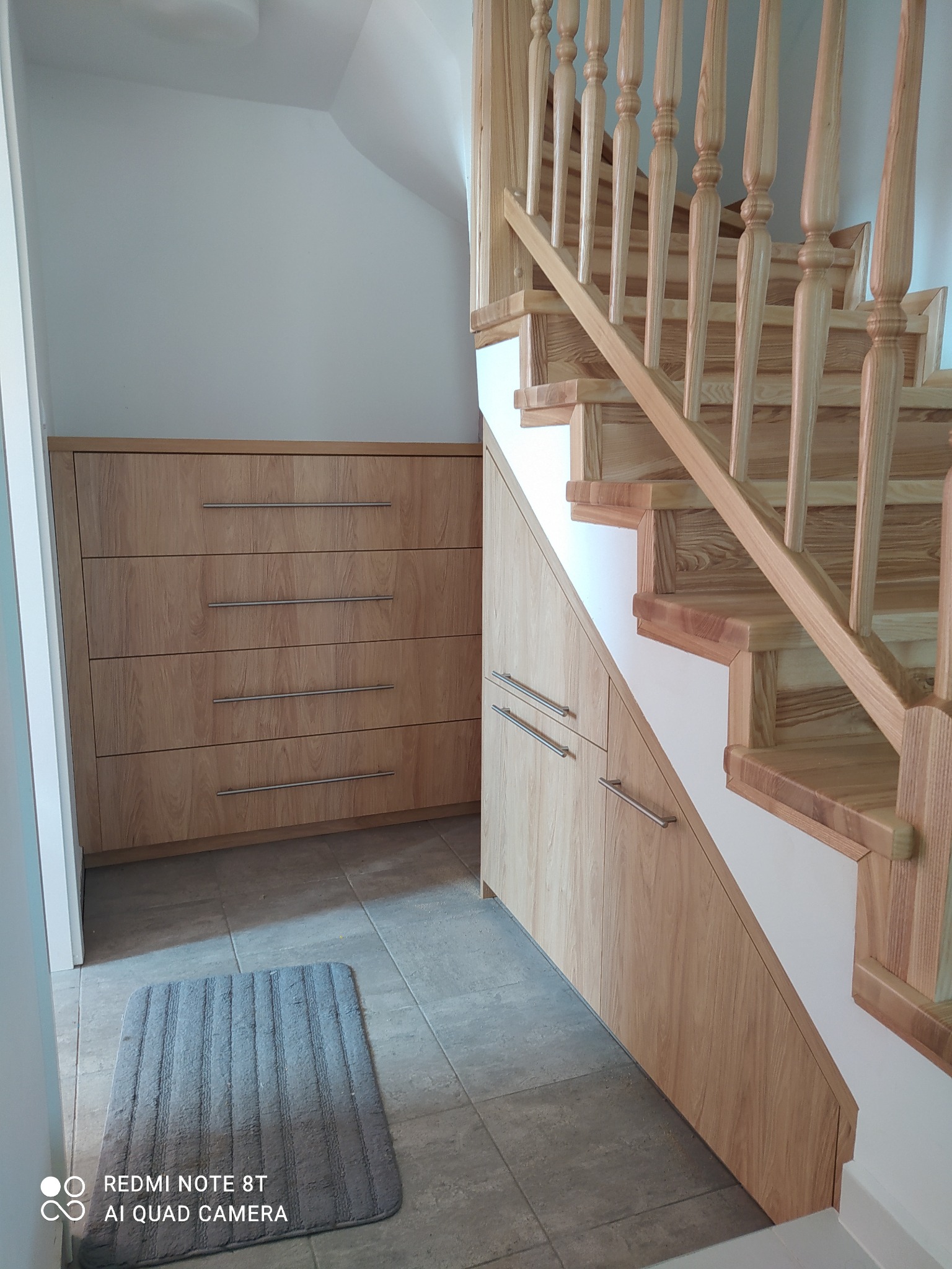 Drewniane schody z zabudową szafek pod schodami, obok komoda z szufladami, szary dywanik na szarej podłodze.