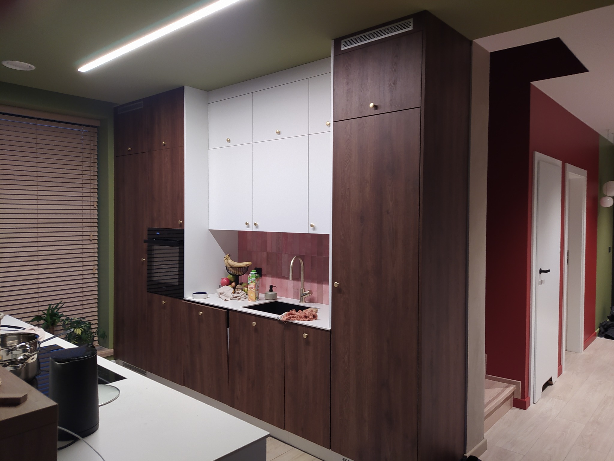 Nowoczesna kuchnia z drewnianymi szafkami dolnymi i białymi górnymi, złotymi gałkami, różowymi płytkami nad blatem i owocami w zlewie.