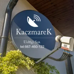 Antena satelitarna zamontowana na ścianie budynku, z widocznym logo firmy 'KaczmareK Usługi Sat.' i numerem telefonu, otoczona pnącą roślinnością.