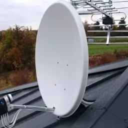 Biała antena satelitarna zamontowana na szarym dachu z widocznymi przewodami i anteną kierunkową w tle na tle jesiennego krajobrazu.