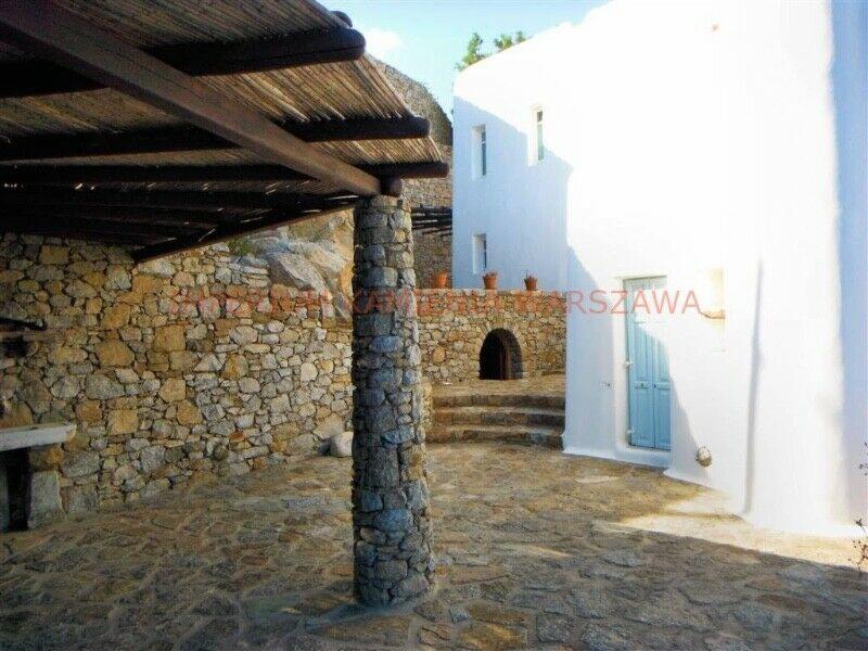 Kamienny taras z zadaszeniem trzcinowym wspartym na kolumnie z kamienia polnego, biały budynek w tle z niebieskimi drzwiami i kamiennymi schodami.