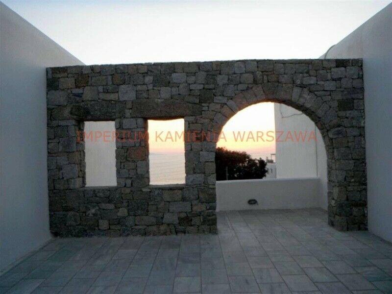 Kamienna ściana z dwoma prostokątnymi otworami i łukowatym przejściem, prowadząca na taras z kamienną posadzką, z widokiem na morze o zachodzie słońca.