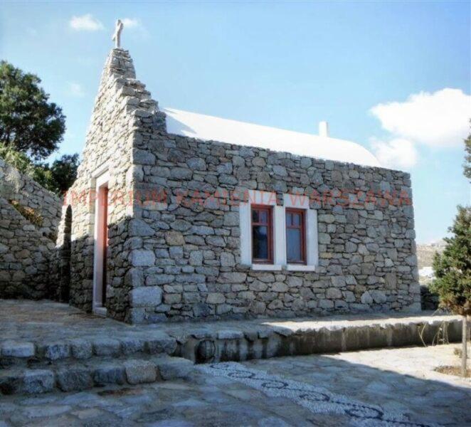 Kamienna kapliczka z białym dachem i krzyżem na szczycie, widoczna z poziomu kamiennego tarasu z dekoracyjnym wzorem z kamyków.