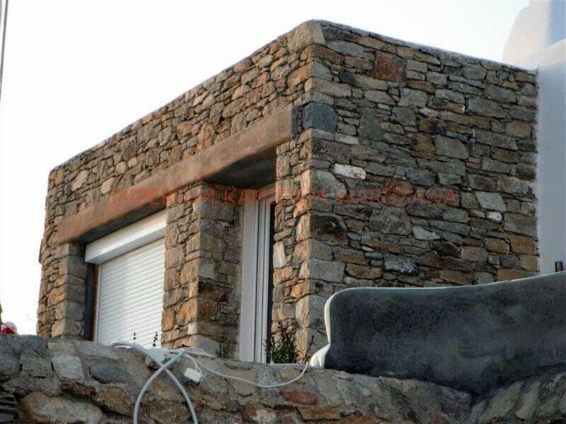 Fragment budynku z elewacją z nieregularnego kamienia, z oknem zabezpieczonym roletą, widoczne kable i fragment tarasu z surowym wykończeniem.