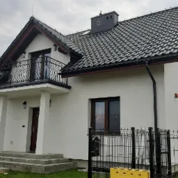 Nowo wybudowany, biały dom jednorodzinny z ciemną dachówką, balkonem z ozdobną balustradą i czarnymi rynnami, ogrodzony metalowym płotem.