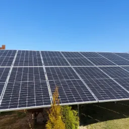 Duży panel słoneczny z widoczną konstrukcją wsporczą, na tle błękitnego nieba i zieleni, z fragmentem budynku w tle.