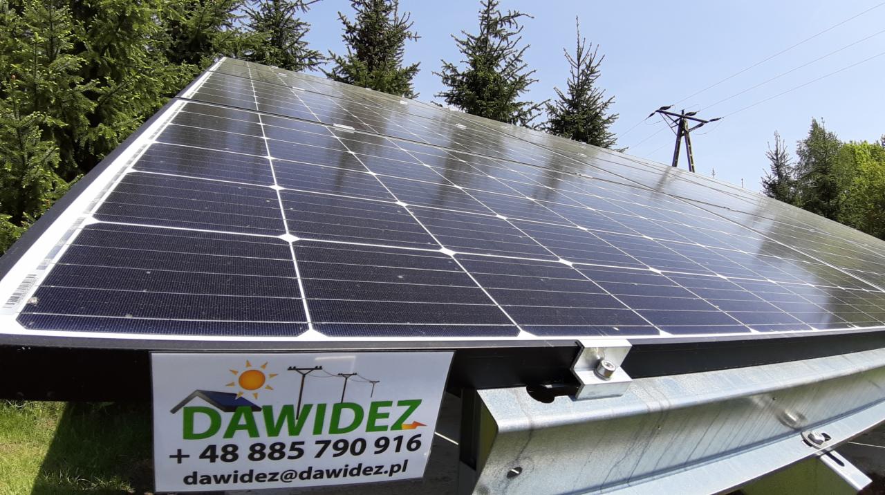 Panel słoneczny zamontowany na metalowej konstrukcji, z widocznym logo firmy DAWIDEZ i numerem telefonu, na tle drzew i słupa energetycznego.
