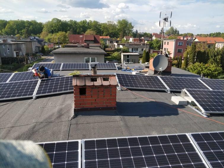Montaż paneli słonecznych na dachu pokrytym papą, widoczny komin z cegły, antena satelitarna i pracownik w niebieskiej koszulce.