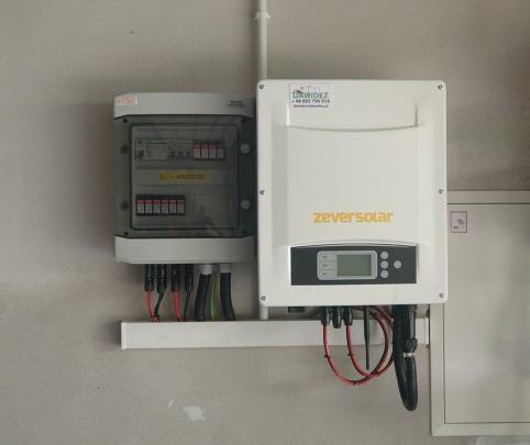 Zamontowany inwerter Zeversolar obok skrzynki z bezpiecznikami, kable poprowadzone wzdłuż ściany na białej listwie.