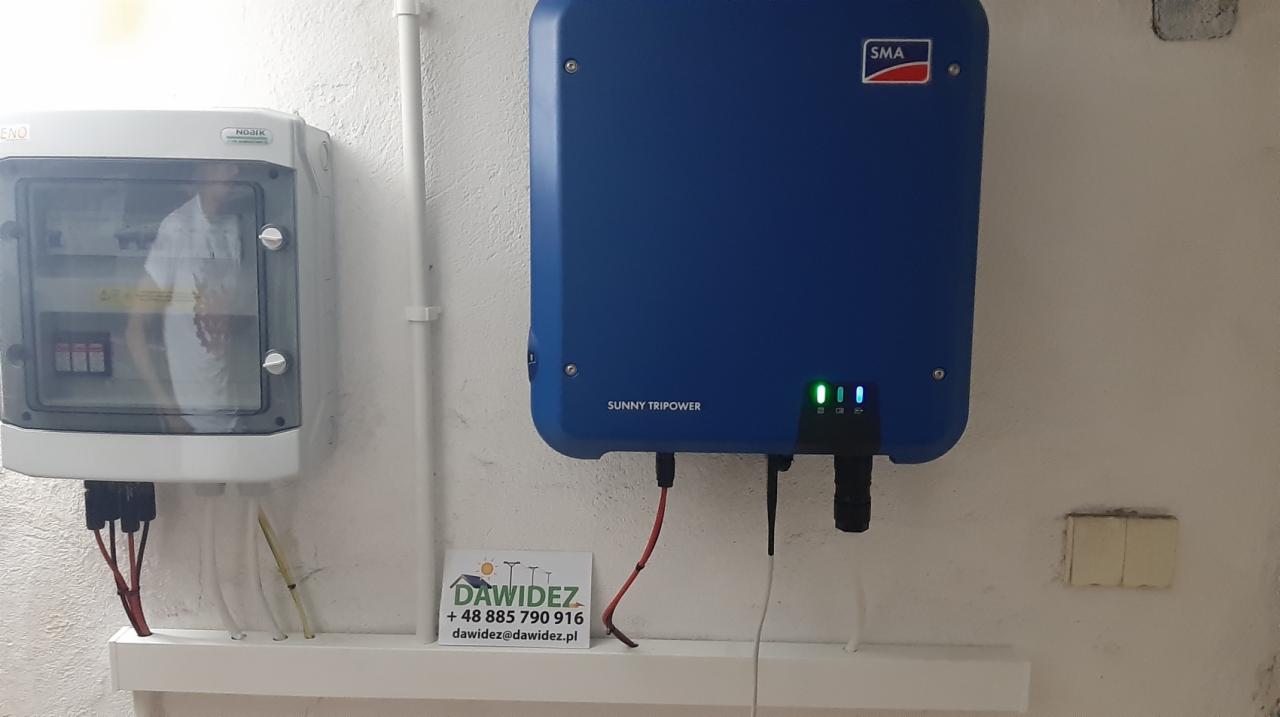Niebieski inwerter Sunny Tripower firmy SMA zamontowany na ścianie obok skrzynki elektrycznej z widocznymi przewodami i wizytówką firmy.
