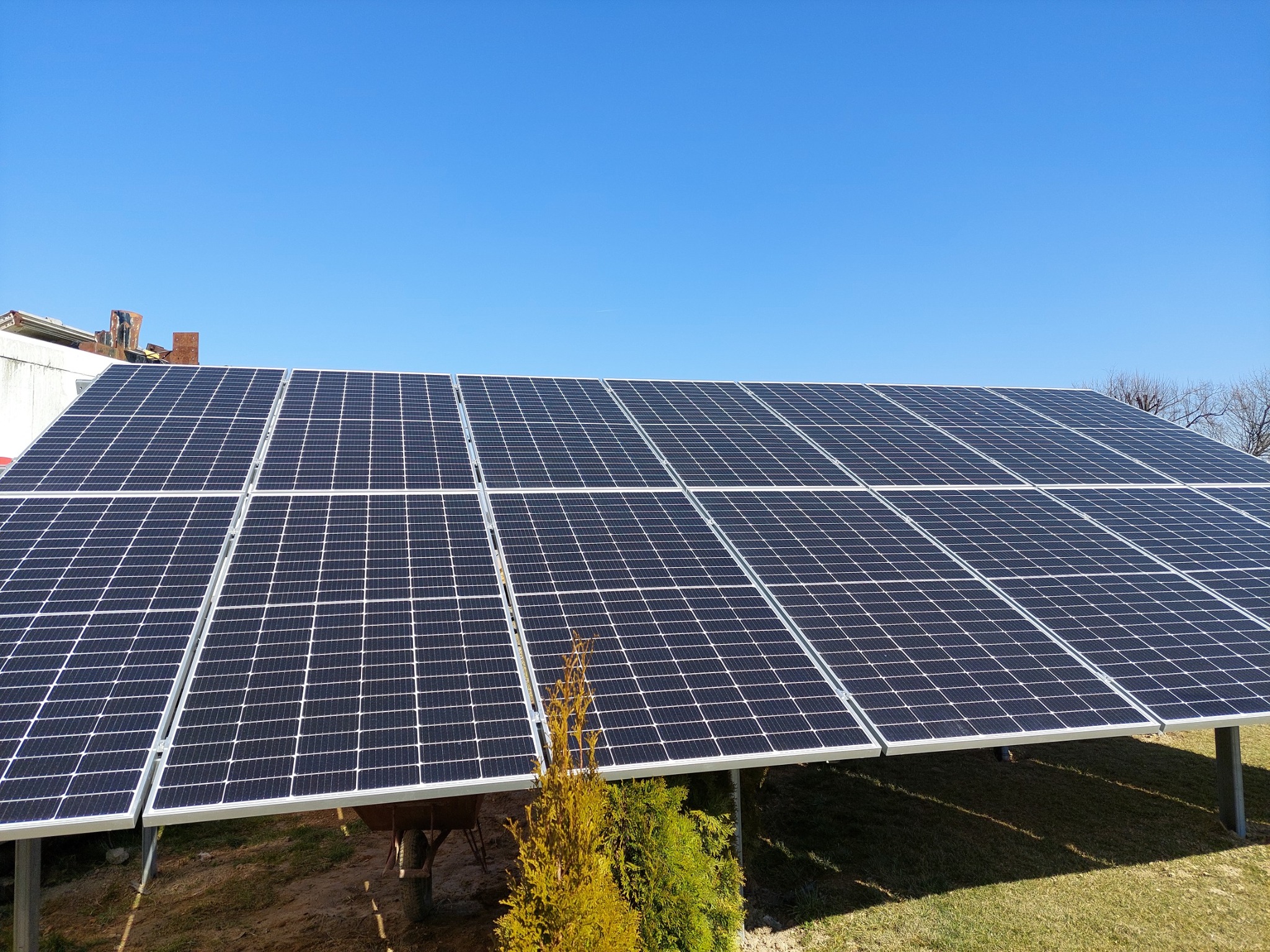 Duży panel słoneczny z widoczną konstrukcją wsporczą, na tle błękitnego nieba i zieleni, z fragmentem budynku w tle.