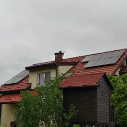 Dom jednorodzinny z panelami fotowoltaicznymi na czerwonym dachu, widok zza brązowego ogrodzenia i zielonych krzewów w pochmurny dzień.