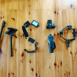 Sprzęt wideofilmowy ułożony na drewnianej podłodze tworzący napis 'Vision': dwa statywy, mikrofon, kamera, gimbal, pasek do kamery Lumix i inne akcesoria.