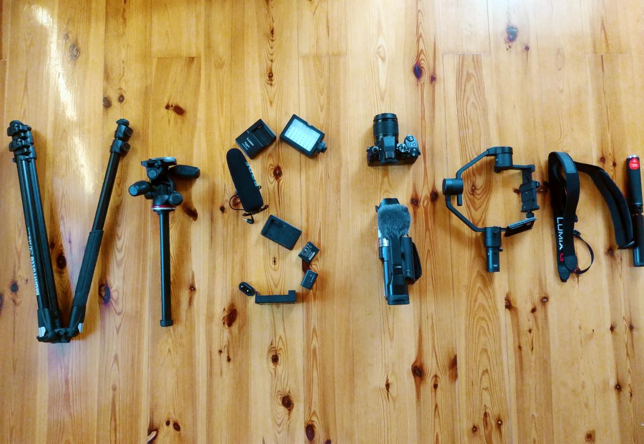 Sprzęt wideofilmowy ułożony na drewnianej podłodze tworzący napis 'Vision': dwa statywy, mikrofon, kamera, gimbal, pasek do kamery Lumix i inne akcesoria.