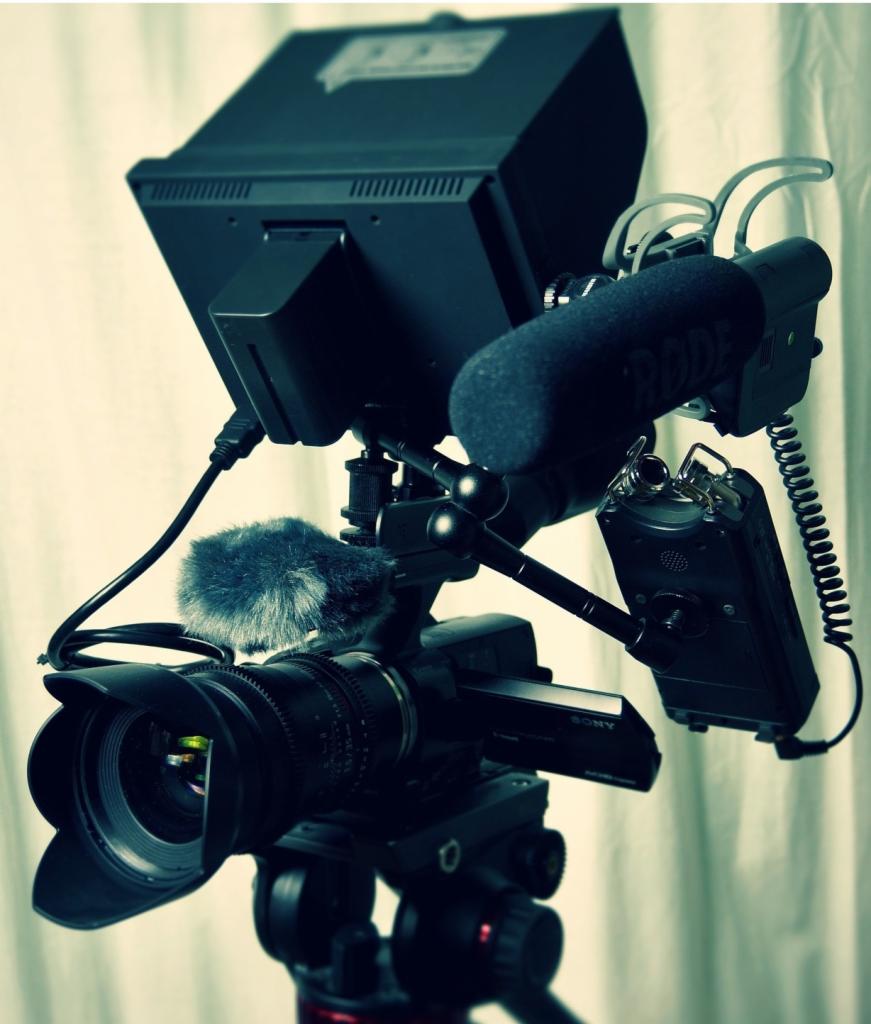 Profesjonalna kamera Sony z mikrofonami typu shotgun i lavalier, zamontowana na statywie, z monitorem podglądowym, ustawiona na tle białej, lekko pogniecionej tkaniny.