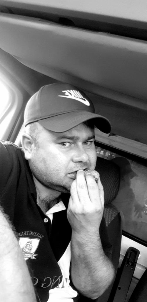 Kierowca w czapce Nike je posiłek w kabinie ciężarówki, prawdopodobnie podczas przerwy w trasie. Zdjęcie czarno-białe, z widocznym oknem i elementami wnętrza pojazdu.