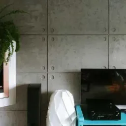 Ściana z płyt betonowych imitujących architektoniczny beton, zdobiona regularnymi okrągłymi wgłębieniami, w salonie z nowoczesnym telewizorem i geometryczną lampą, z wiszącą półką z rośliną.