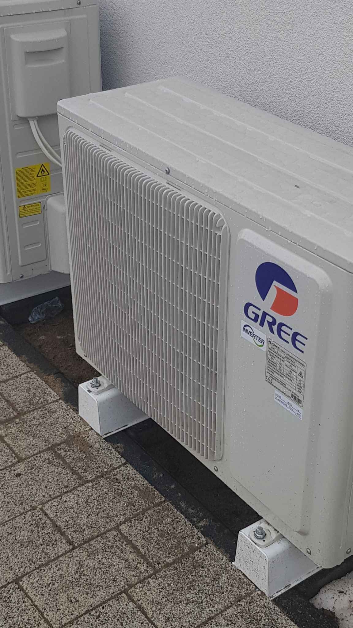 Zewnętrzna jednostka klimatyzacji marki Gree Inverter, zamontowana na betonowych podstawkach, widoczne krople deszczu na obudowie.