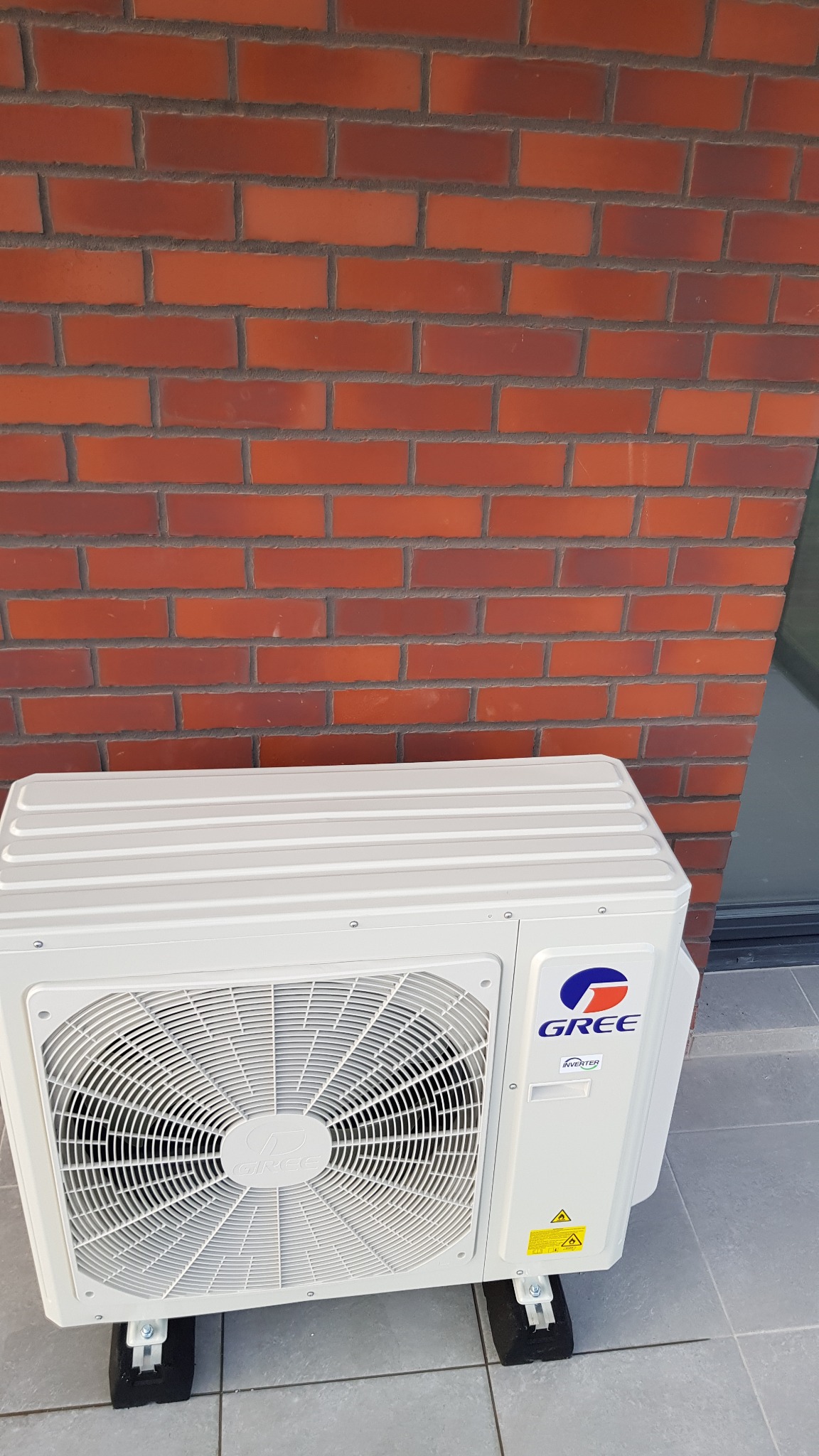 Zewnętrzna jednostka klimatyzacji marki Gree, zamontowana na czarnych podkładkach, na tle ceglanej ściany i szarej posadzki.