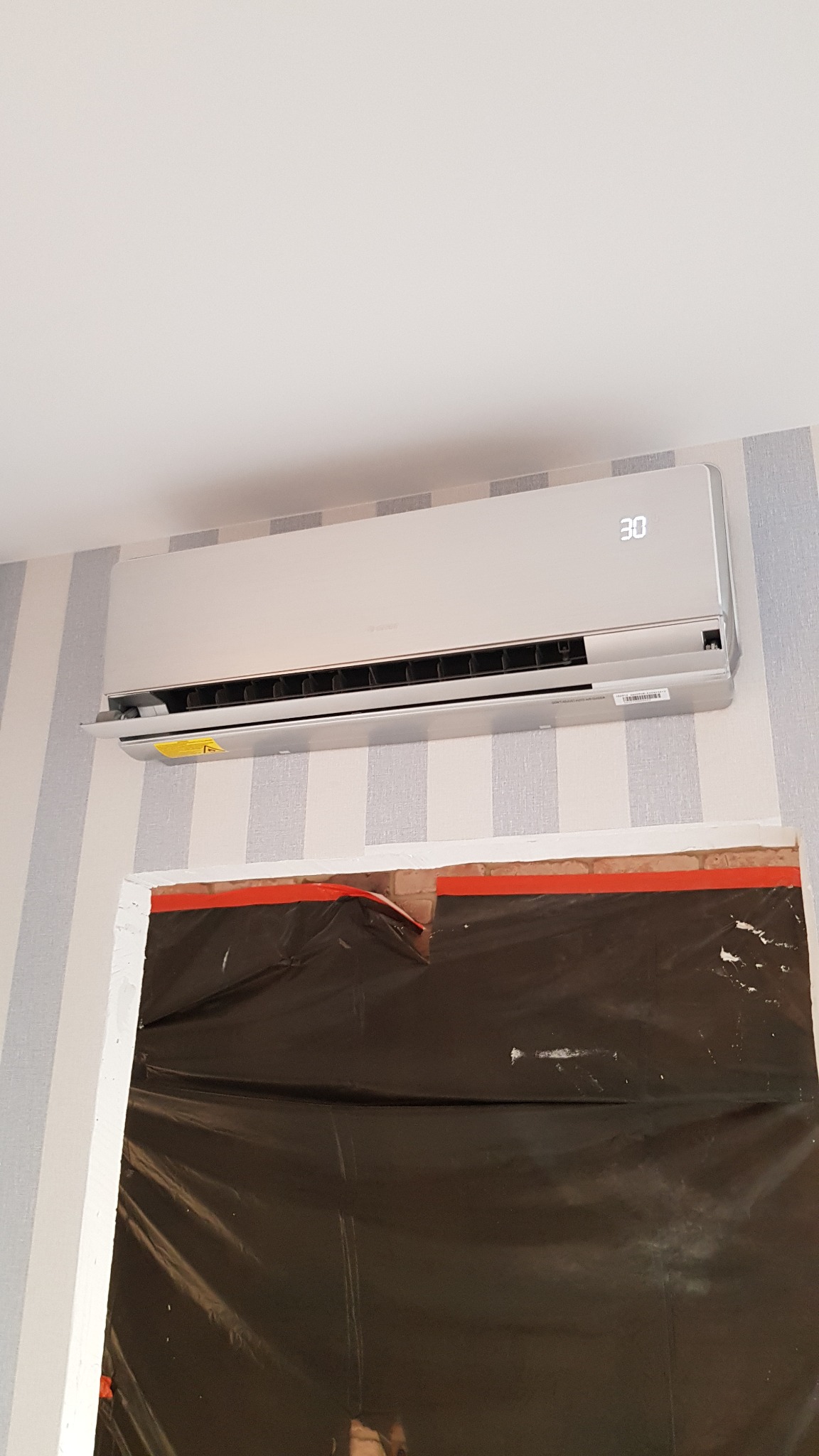 Zamontowany klimatyzator na ścianie w pokoju z tapetą w pionowe pasy, zabezpieczonym folią malarską przed zabrudzeniem.