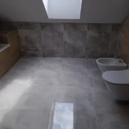 Wykończona łazienka na poddaszu z płytkami imitującymi beton na ścianach i podłodze, drewnopodobnymi płytkami przy wannie oraz oknem dachowym wpuszczającym światło.