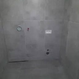 Szare płytki ceramiczne ułożone na ścianach i podłodze łazienki z widocznymi elementami instalacji sanitarnych: odpływ liniowy, przyłącza wodne i stelaż podtynkowy WC.