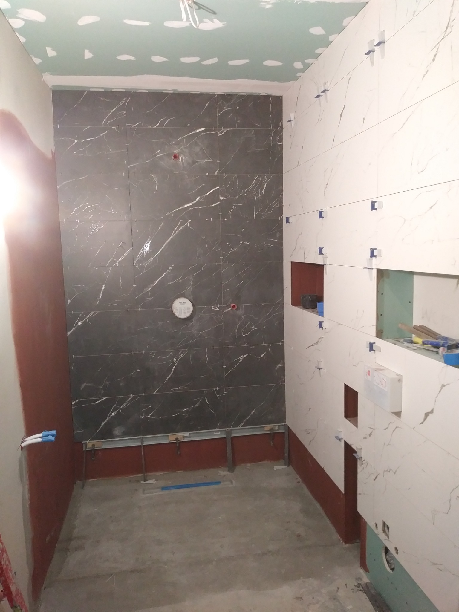 Wnętrze łazienki w trakcie prac wykończeniowych: układanie płytek imitujących marmur w odcieniach szarości i bieli, widoczne listwy montażowe, otwory pod instalacje sanitarne i elektryczne...