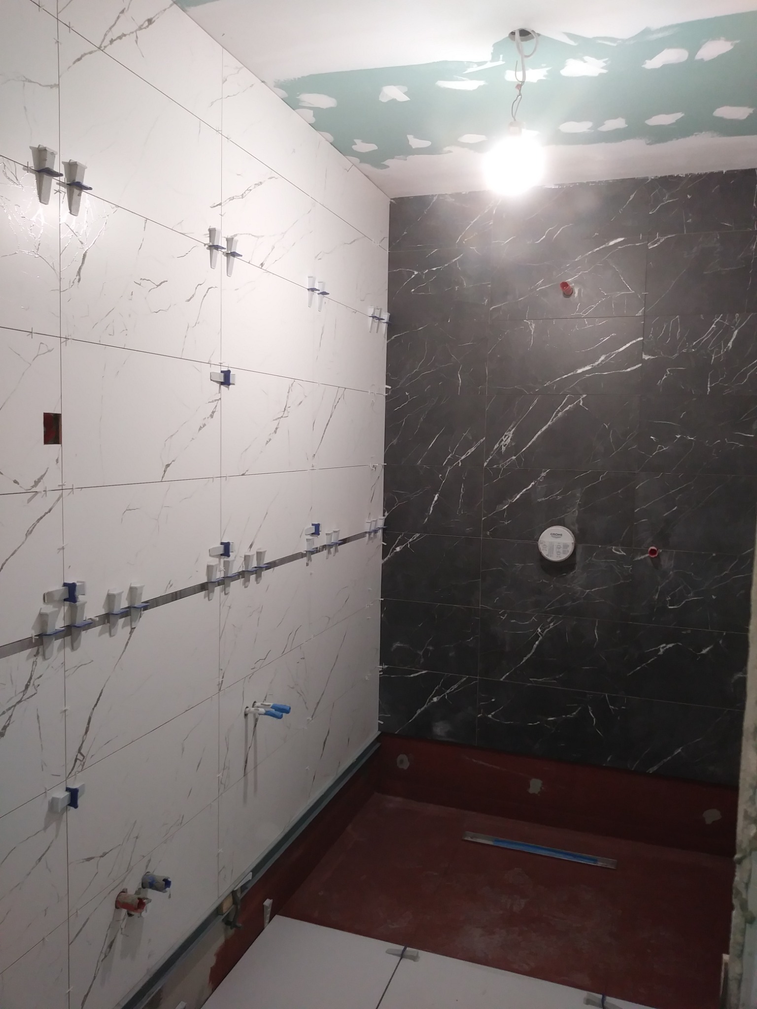 Łazienka w trakcie wykańczania: białe i czarne płytki marmurowe na ścianach, system poziomowania płytek z krzyżykami, przygotowana podłoga pod prysznic, surowy sufit z widocznymi łączeniami płyt...