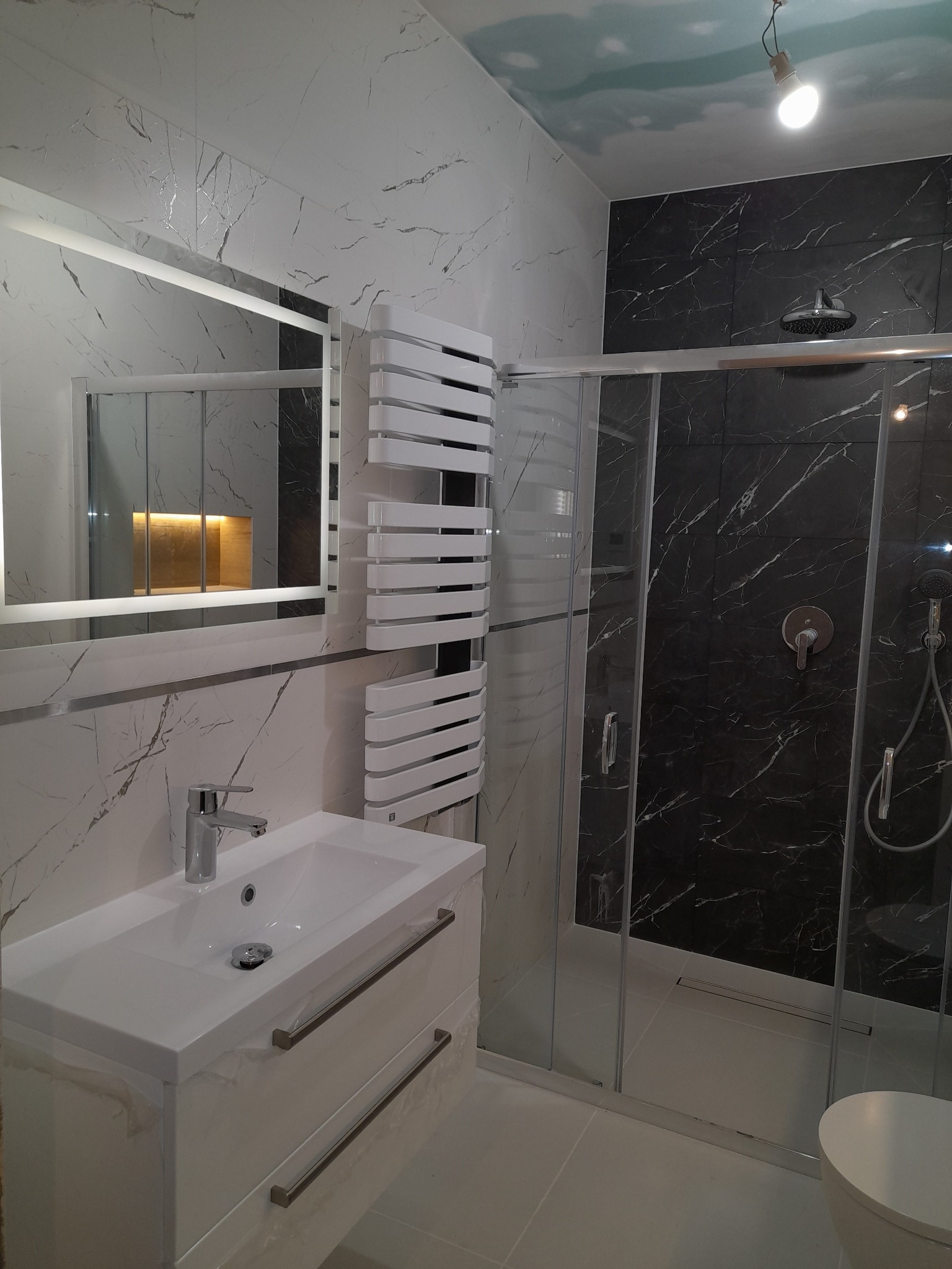 Nowoczesna łazienka z białymi marmurkowymi płytkami, czarną kabiną prysznicową typu walk-in, podświetlanym lustrem i grzejnikiem drabinkowym; surowy strop z widoczną żarówką.