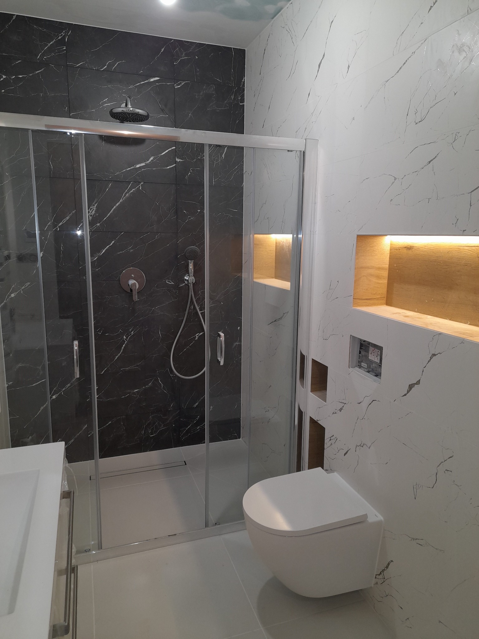 Nowoczesna łazienka z kabiną prysznicową wyłożoną czarnymi płytkami imitującymi marmur, białe płytki marmurowe na ścianie z wnękami oświetlonymi ciepłym światłem, wisząca toaleta.