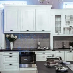 Elegancka, biała kuchnia na wymiar w stylu klasycznym z czarnym blatem, przeszklonymi szafkami, dekoracyjnym panelem nad blatem i ciastem pod kloszem na stole.