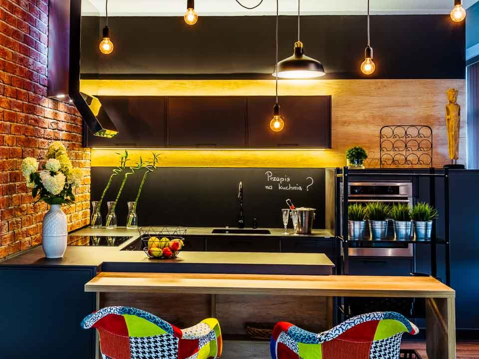 Nowoczesna kuchnia z ceglaną ścianą, czarnymi frontami szafek i drewnianym blatem, oświetlona dekoracyjnymi żarówkami, z wyspą i krzesłami patchworkowymi.