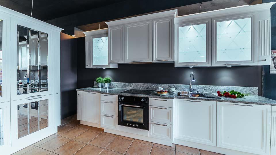 Elegancka, biała kuchnia z frezowanymi frontami, szarym blatem i przeszklonymi szafkami wiszącymi z podświetleniem, uzupełniona o lustrzaną szafę i czarny piekarnik.