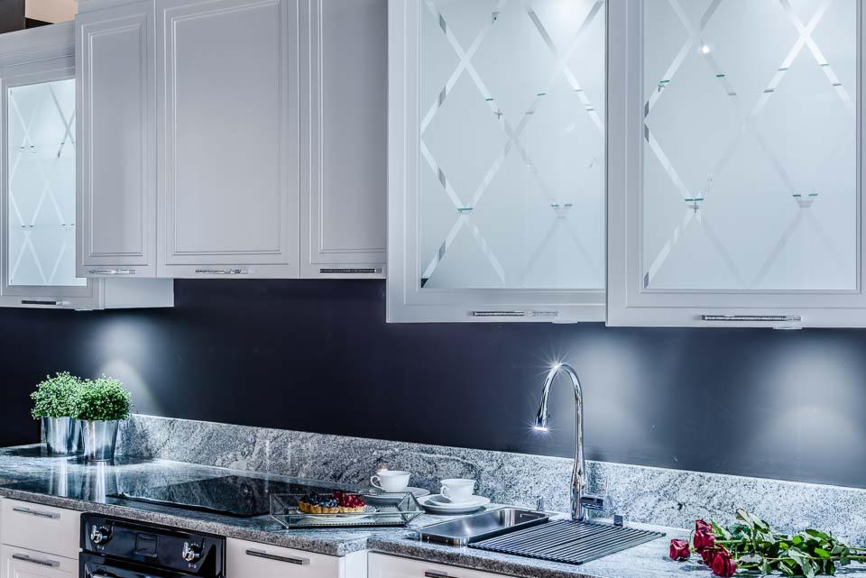 Biała kuchnia z górnymi szafkami z matowym szkłem w romby, szary granitowy blat z kwiatami i ciastem, chromowana bateria.
