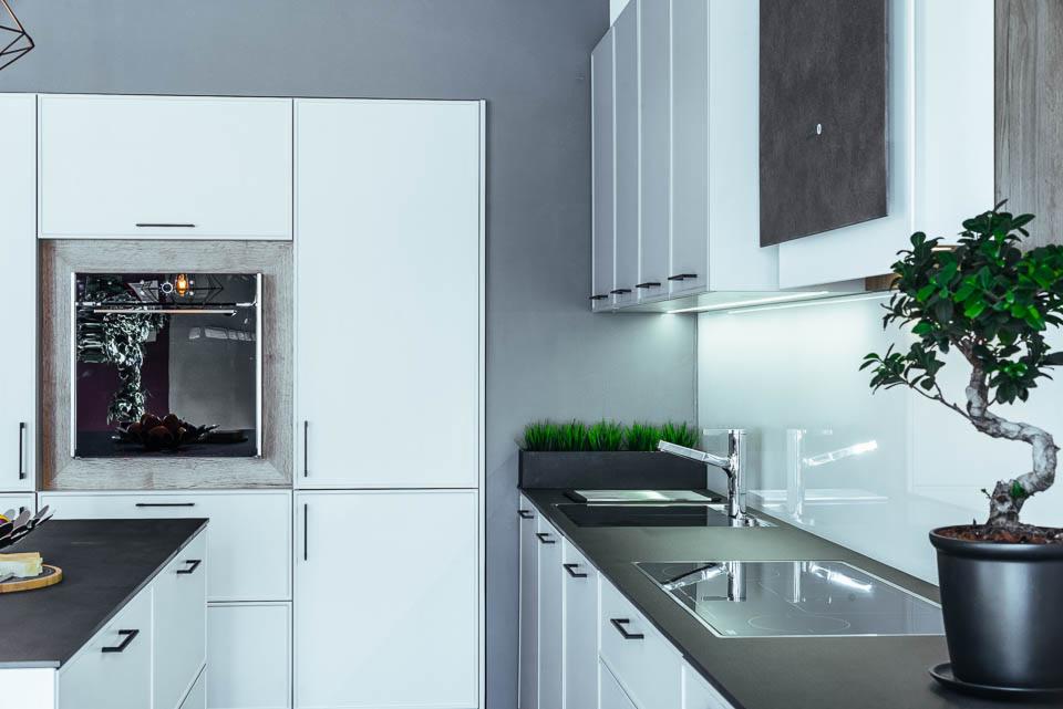 Nowoczesna kuchnia z białymi frontami, czarnymi blatami i minimalistycznymi uchwytami, uzupełniona o dekoracyjne elementy: bonsai w doniczce i sztuczną trawę w czarnej skrzynce.