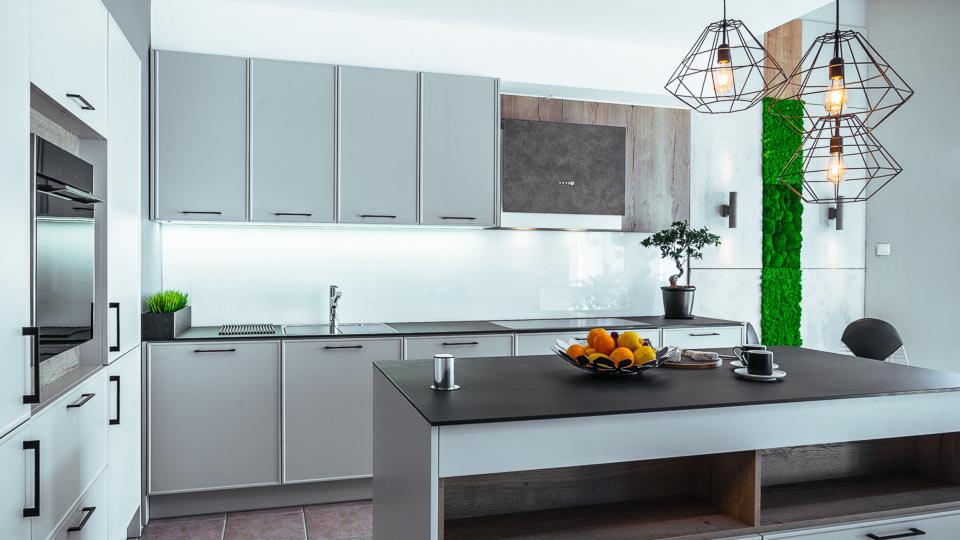 Nowoczesna kuchnia z szarymi szafkami, czarnym blatem wyspy, geometrycznymi lampami wiszącymi i panelem z mchu na ścianie.