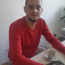 Młody mężczyzna w czerwonej bluzie siedzi przy stole z filiżanką kawy i telefonem. Proste, białe tło.