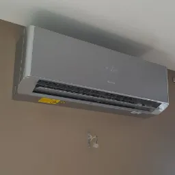 Szara jednostka klimatyzacyjna Gree zamontowana na brązowej ścianie, widoczne przewody elektryczne w miejscu montażu, ustawiona temperatura 16 stopni.