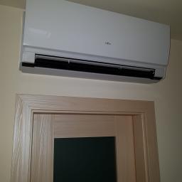 Klima Control - Biała jednostka klimatyzacyjna marki Fujitsu zamontowana na ścianie nad drewnianymi drzwiami z czarnym wypełnieniem