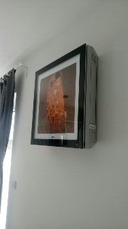 Klimatyzator ścienny LG Art Cool Gallery z obrazem 'Pocałunek' Gustava Klimta w czarnej ramie, zamontowany na białej ścianie w jasnym pomieszczeniu, obok zasłony w odcieniach szarości.