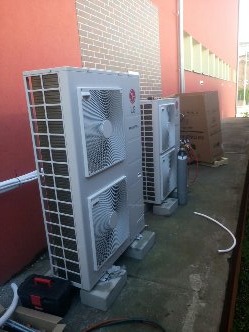 Zewnętrzne jednostki klimatyzacyjne LG Inverter na betonowych podstawach, podczas instalacji na tle czerwonej ściany budynku. Widoczne butle z gazem chłodniczym i skrzynka narzędziowa.
