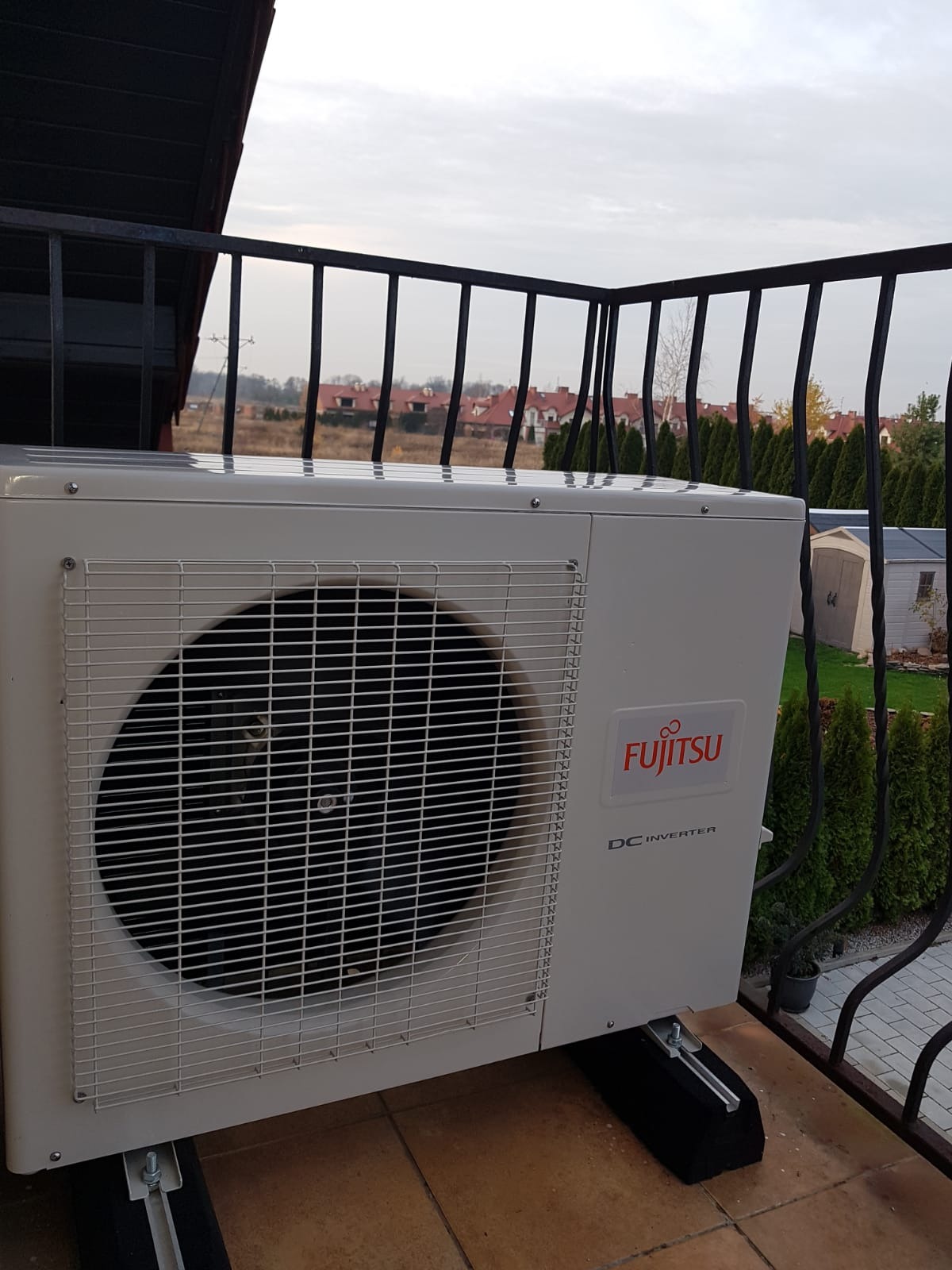 Zewnętrzna jednostka klimatyzacji Fujitsu DC Inverter zamontowana na balkonie z widokiem na osiedle domów i zielone tuje.
