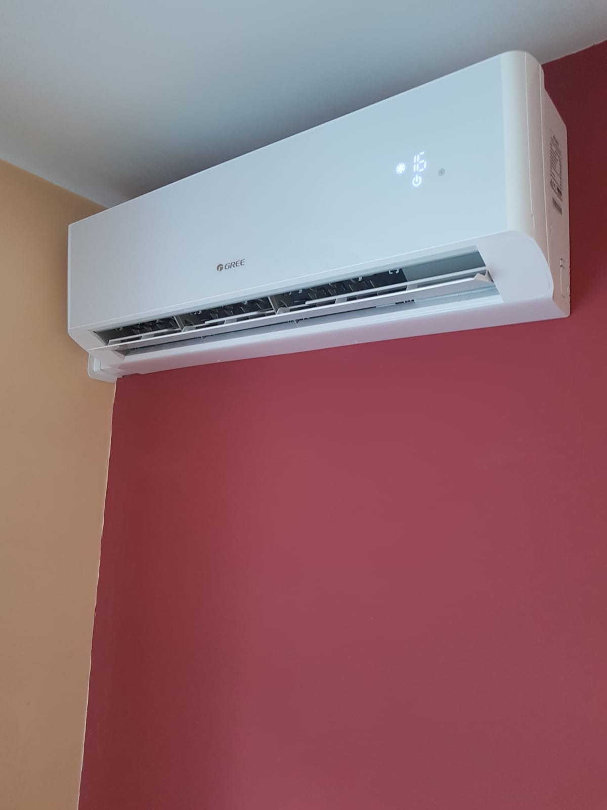 Biała klimatyzacja marki Gree zamontowana na ścianie pomalowanej na kolor bordowy i beżowy, widoczny wyświetlacz z temperaturą 16 stopni.
