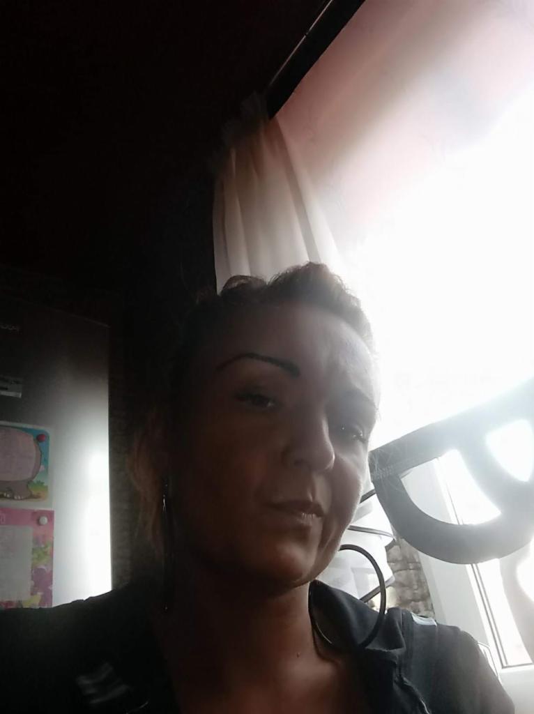 Portret kobiety z ciemnymi włosami, stojącej w słonecznym pokoju z widoczną lodówką z magnesami w tle.