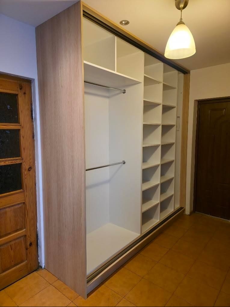 Jasna szafa wnękowa z przesuwnymi drzwiami z płyty drewnopodobnej, z półkami i drążkami na ubrania, widoczna w korytarzu.
