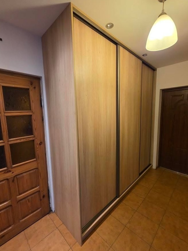 Jasna szafa wnękowa z przesuwnymi drzwiami, dopasowana do wnętrza przedpokoju z brązową podłogą i drewnianymi drzwiami. Widoczna lampa sufitowa.