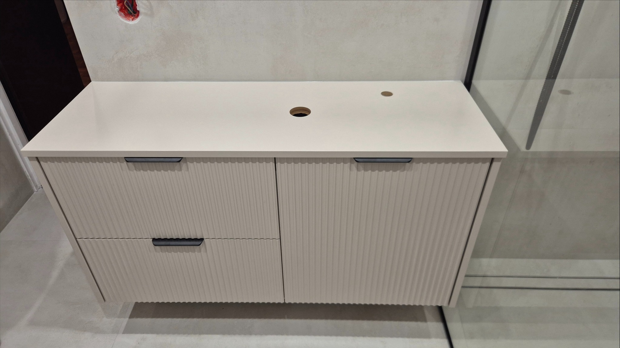 Beżowa szafka łazienkowa z frezowanymi frontami i czarnymi uchwytami, widoczna z bliska, z otworem na umywalkę i baterię. Obok kabina prysznicowa.