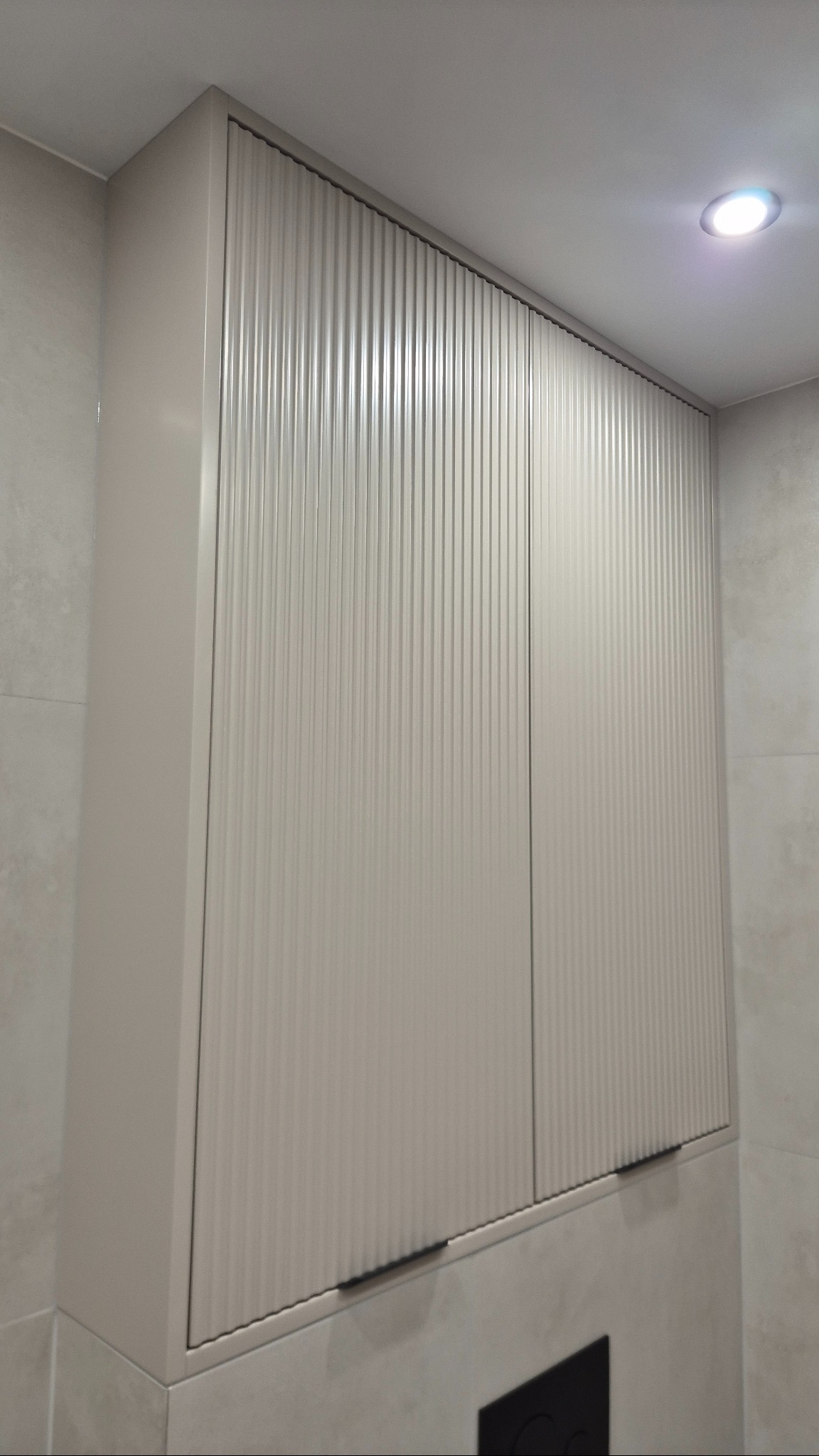 Beżowa szafka łazienkowa z ryflowanymi drzwiami, zamontowana na ścianie z jasnych płytek. Minimalistyczny design, oświetlenie sufitowe.