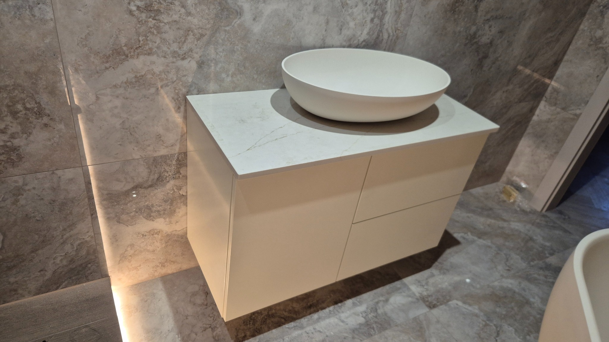 Biała szafka łazienkowa z umywalką nablatową na tle marmurowej ściany i podłogi. Minimalistyczny design, jasne kolory, nowoczesne wykończenie.